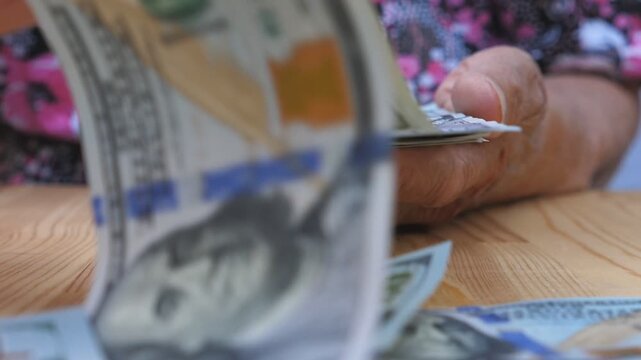 Grandmother holding bundle of money in hands and scattering foreign currency on the desk. Arms of old woman throwing one hundred dollar bills on the table in front to camera. Close up Slow motion