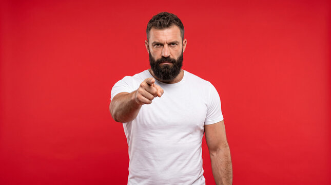 Angry bearded man in white t-shirt pointing finger forward against a red background