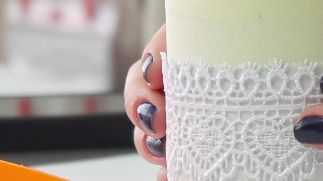 A close-up view of a person's hands, adorned with dark, glossy nail polish, delicately holding a cream-colored candle.