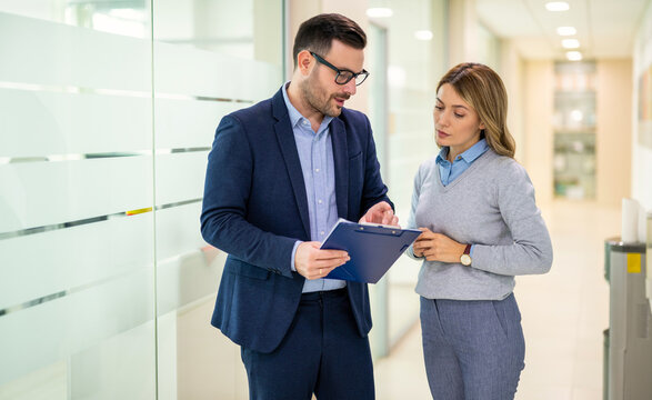 Business colleagues standing in an office hallway, reviewing documents on a clipboard and discussing work details during a professional conversation.