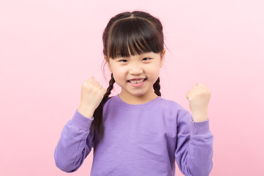 Excited little Asian girl raising fists in victory celebrating success on a pink background.