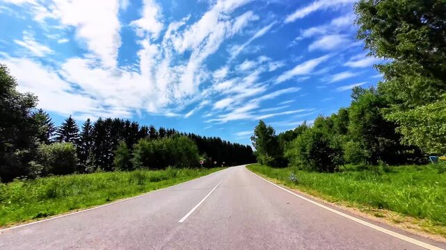 Winding asphalt road bordered by lush green trees and vibrant blue sky full of clouds