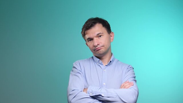 Man with dark hair and blue shirt on a teal background expressing feelings of sadness, disapproval, boredom, and doubt, showing skepticism with crossed arms