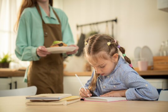 Caucasian little girl doing homework at kitchen table while mother bringing snack. Cute focused daughter writing in notebook with mother holding a sandwich plate in background.