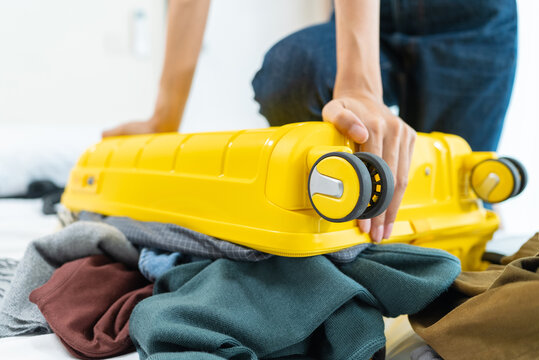 Close-up of Asian woman sitting on an overpacked yellow suitcase to close it. Concept of overpacking issues, travel preparation struggles and holiday planning.