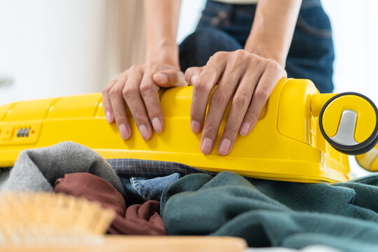 Close-up of Asian woman sitting on an overpacked yellow suitcase to close it. Concept of overpacking issues, travel preparation struggles and holiday planning.