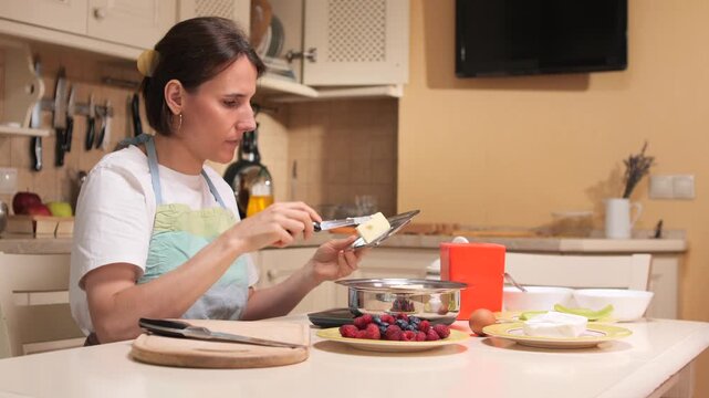 Young woman preparing a dessert in a home kitchen, grating butter into a bowl while having fresh berries and other ingredients ready on the table