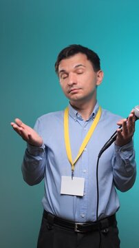 Vertical video man looking confused and shrugging with open hands while holding a microphone and wearing an empty yellow lanyard and id badge, standing against a blue background