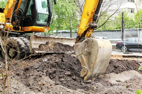 Yellow excavator loader cabin heavy bucket arm and front dozer blade mechanism moving ground soil dirt pile at urban road infrastructure construction work repair site area daytime background
