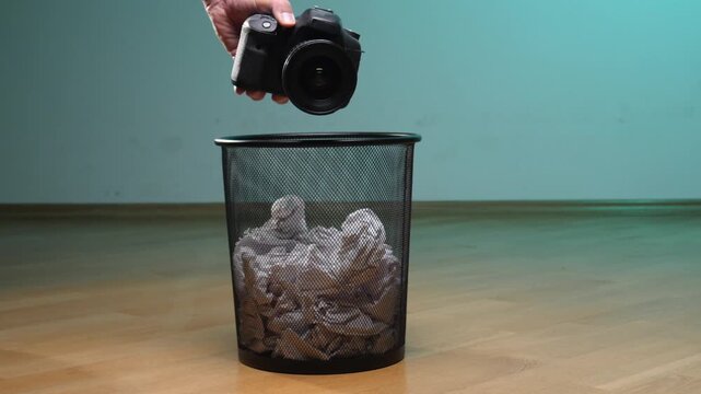 Human hand dropping an outdated professional digital single lens reflex camera into a metal trash bin filled with crumpled waste paper, symbolizing obsolescence and electronic waste disposal