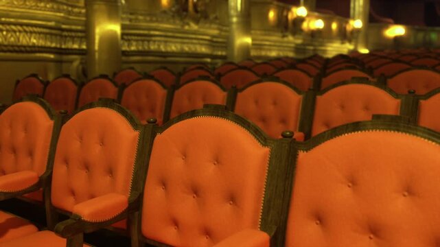 Red velvet theater seats ornate auditorium, empty rows facing dimly lit proscenium, plush upholstery, brass fixtures, patterned carpet, intimate golden