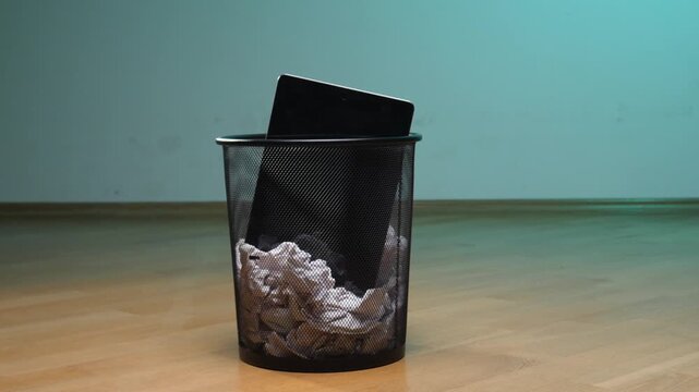 Person disposing of an obsolete tablet computer into a black mesh trash can filled with crumpled paper, symbolizing digital detox, electronic waste, and technological obsolescence