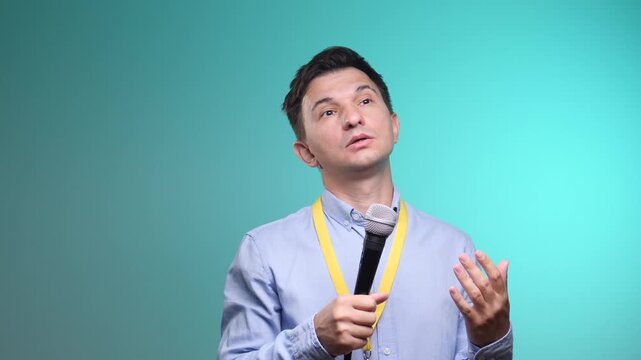 Man speaker wearing a light blue shirt and yellow lanyard, holding a microphone, and gesturing while delivering a speech or presentation against a blue background