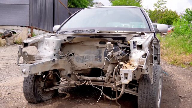 Front view of a wrecked silver car in a junkyard, heavily dismantled with the engine, bumper, and headlights missing, showing exposed chassis, wiring, and a flat front tire
