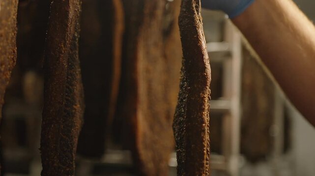 Gloved hand inspects thick seasoned biltong slabs hanging on curing racks in a dim meat drying room.