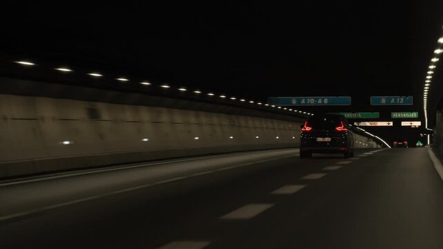 Several automobiles drive through an underground highway tunnel past electronic toll collection signs and route directions in France. Metropolitan road infrastructure transit commuter voyage.