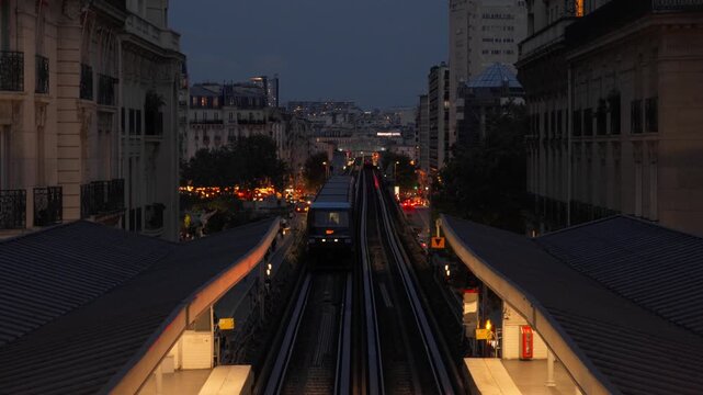 A passenger metro train arrives at an elevated station along urban tracks at dusk in Paris France. Metropolitan public rail transit infrastructure, urban commuter lifestyle evening voyage.