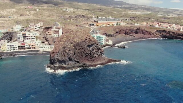 Scenic aerial view flying around the volcanic sea stack of Los Roques de Fasnia and the adjacent coastal village in Tenerife, Canary Islands
