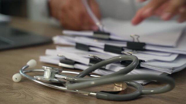 medical paperwork beside a laptop and stethoscope on a wooden desk, documenting information on stacked forms for an organized healthcare records workflow.