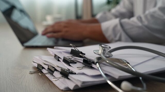 medical paperwork beside a laptop and stethoscope on a wooden desk, documenting information on stacked forms for an organized healthcare records workflow.