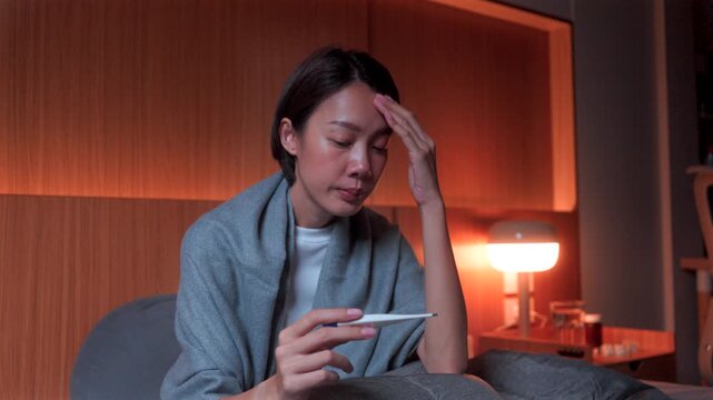 Exhausted woman with a headache reading a thermometer in a dark bedroom with medicine nearby. Concept of feverish symptoms, influenza, and medical self-care at home.