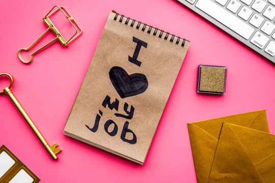 Happy at work. Handwritten motto I love my job in notebook near stationery on pink background top view