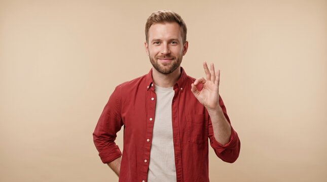 Smiling man with short brown hair wearing a red button down shirt over a white t shirt making an ok hand gesture against a solid light beige background