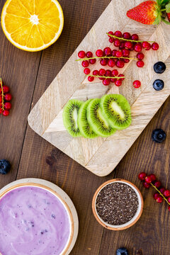 Cook trendy breakfast. Acai smoothie bowl near cutting board with fresh fruits, berries, chia seeds on dark wooden background top view
