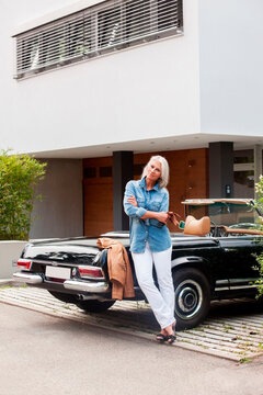 Mature woman leaning on convertible car in front of her one-family house