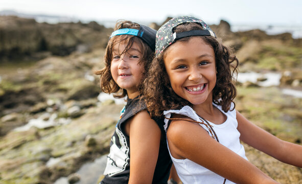 Spain, Gijon, portrait of smiling little girl and her friend sitting back to back at rocky coast