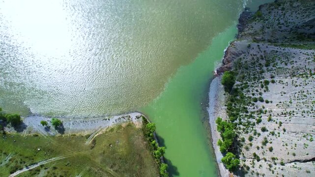 Aerial river confluence showing turquoise mountain stream meeting muddier brown-green channel along rocky shoreline under strong midday sun
