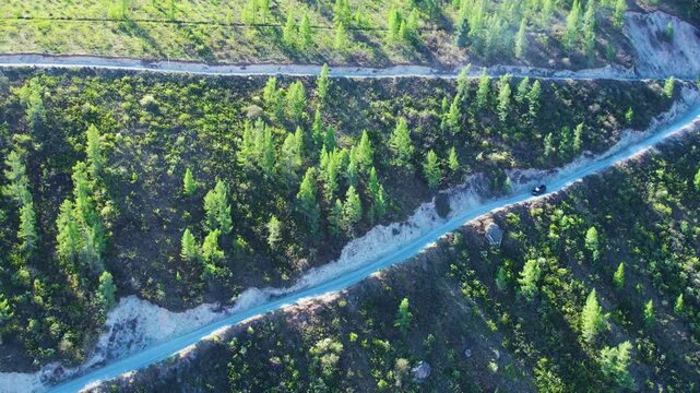 Diagonal aerial view of narrow dirt mountain road with lone vehicle descending switchback through green conifer hillside under bright sunlight and deep shadow, with road, dirt, mountain, forest detail