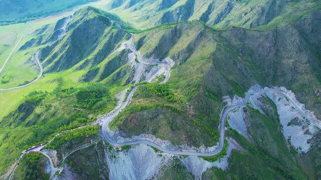 Aerial view of winding mountain pass with steep ridgelines, rocky cliffs, green slopes, hairpin curves, and scenic roadside turnout across rugged highland landscape