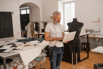 A designer works on a laptop in a bright studio surrounded by materials and tools for creation. © LIGHTFIELD STUDIOS