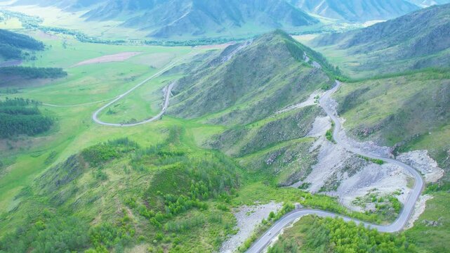 Aerial view showing winding mountain road across layered ridgelines and green valley under sunset light, rocky slopes, distant peaks, soft haze, wide scenic panorama