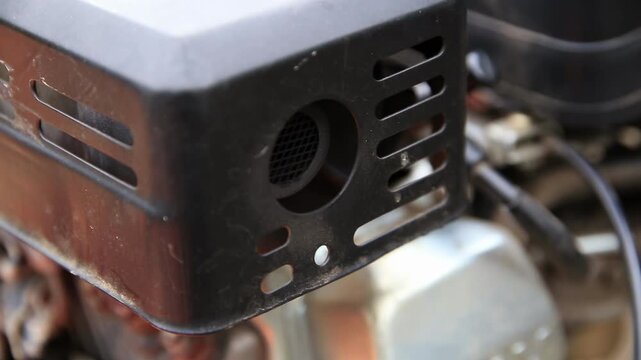 Close up view of dirty black metal protective cover on engine machinery with ventilation slots and mesh screen showing details of exhaust or air intake component and wear
