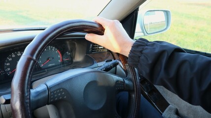 view from inside the car: a driver in a dark windbreaker holds the top of the steering wheel. The speedometer, tachometer, and air vent are visible, and the roadside is reflected in the outside mirror © Skyliz