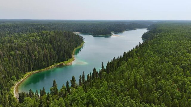 Drone glides above lush forest alongside the vibrant green waters of Slim Lake, showcasing the pristine wilderness of Northern Ontario, Canada.