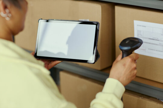 Close up of young adult woman scanning box label with barcode reader while checking tablet in warehouse, managing inventory and order tracking. Ideal for logistics and ecommerce content