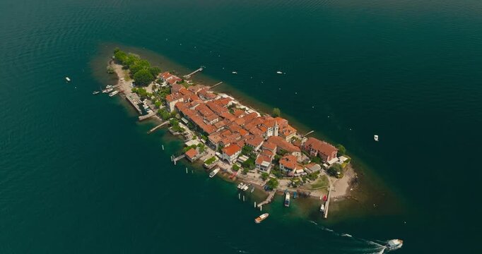 Drone flies over Isola dei Pescatori, Borromean Island, on Lake Maggiore, revealing orange rooftops, deep blue water and a boat passing along the shoreline in a beautiful Italian summer landscape.