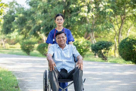 Elderly man patient in a wheelchair being cared for by a medical professional. Asian nurse take care senior male sit on wheelchair in park, outdoor.