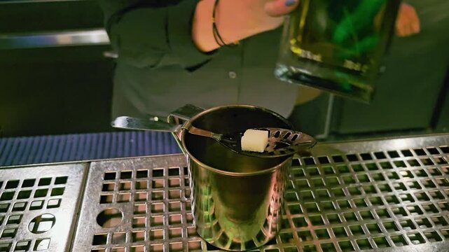 Traditional absinthe ritual at a dim bar, featuring sugar cube on slotted spoon, metal cup, green bottle, bartender tools, and reflective stainless surface under colorful lighting.