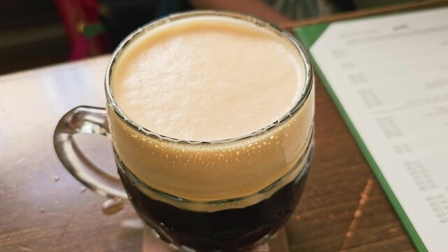 Close-up of a frothy dark beer mug on a wooden pub table, with creamy foam, chilled glass, condensation drops, blurred menu, and warm restaurant atmosphere for craft beer stock videos.