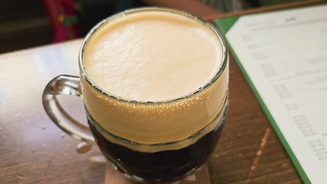 Close view of a beer mug being picked up in a bar, with creamy foam, dark lager, wet glass texture, wooden tabletop, menu in background, and relaxed European pub setting at night.