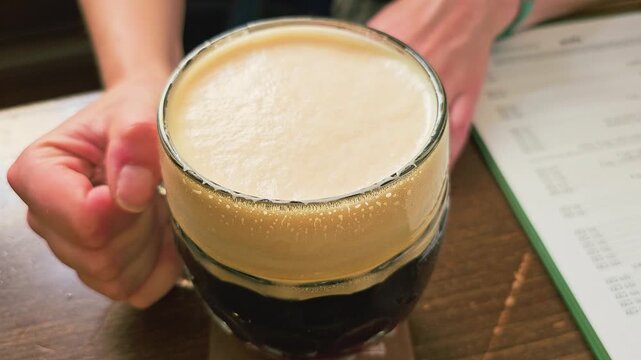 Close-up of a hand lifting a frothy dark beer mug from a wooden pub table, with creamy foam, chilled glass, blurred menu, and warm bar atmosphere for restaurant drinks and nightlife.