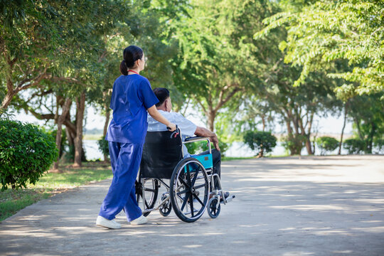 Female nurse pushing elderly man jn wheelchair outdoors serene environment. Image conveys compassionate elderly care and assisted living in a natural setting.