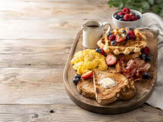 Fototapeta premium Rustic breakfast platter with fluffy waffles fresh berries scrambled eggs bacon toast butter and maple syrup served on a wooden board beside coffee and fruit in a warm farmhouse table setting