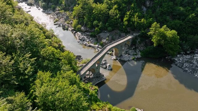 Devils bridge Dyavolski most high angle drone shot shadow reflection water surface green forest river valley curve summer midday sunshine historical tourism concept Bulgaria.