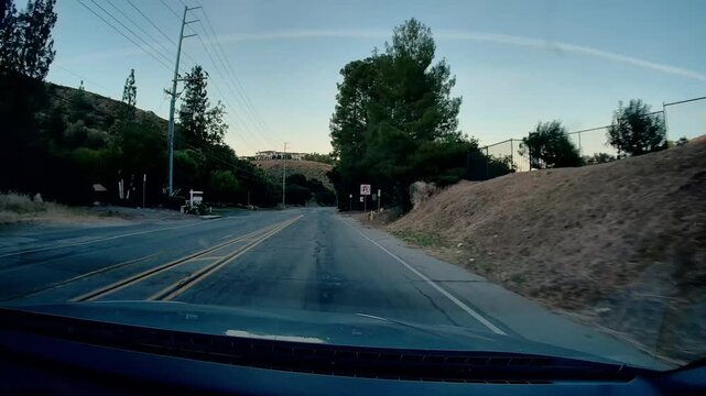 A dashcam view of Mulholland Hwy. driving along scenic winding road, surrounding hills with dense vegetation, utility poles, residential structures background, expansive clear sky
