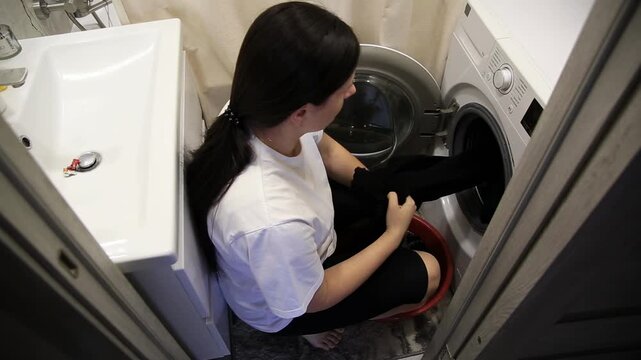 Woman loads black laundry into washing machine from red basin. Domestic chore of cleaning clothes performed indoors bathroom utility area. Daily routine household task.